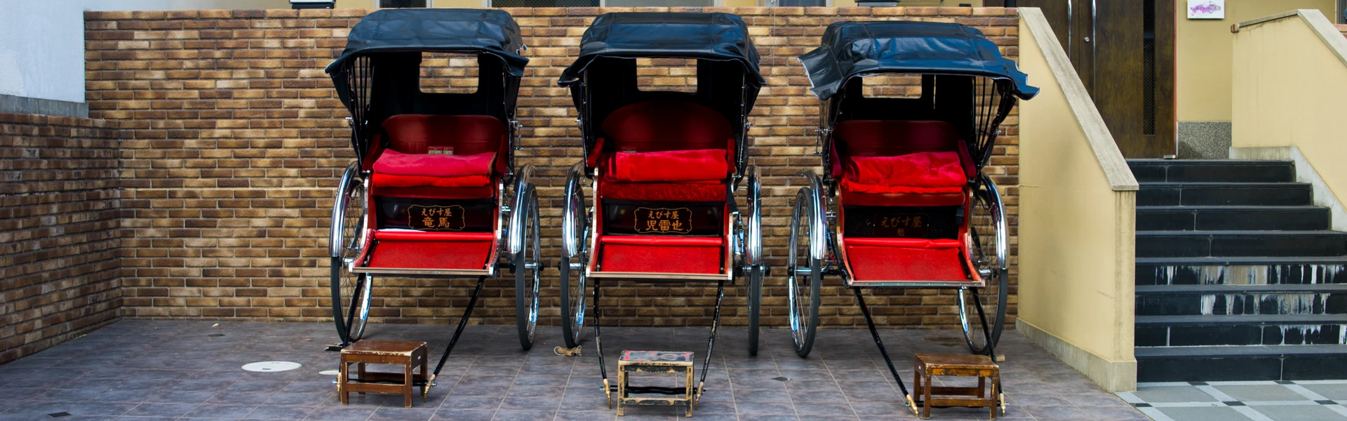 Three red-seated rickshaws parked in a row against a brick wall beside a concrete staircase.