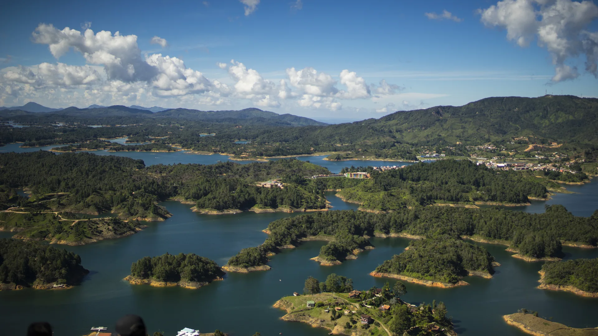 Elevated view over a many-armed reservoir winding between forested green hills, with scattered buildings, small clearings and distant mountains under towering white clouds.