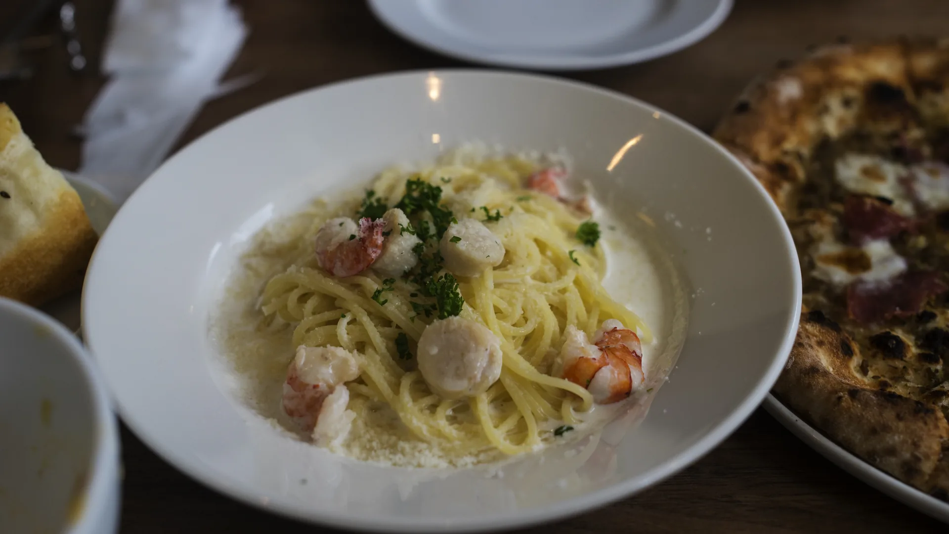 A shallow bowl of thin pasta in a pale cream sauce with shrimp, scallops and chopped parsley, bread and a pizza alongside on a wooden table.