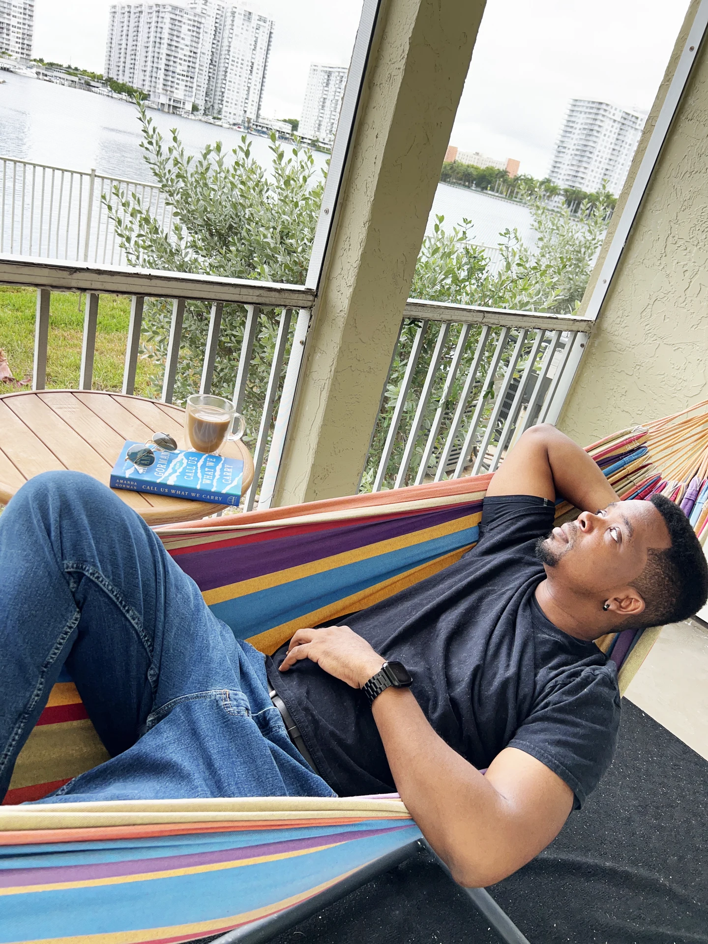 Willie Grady reclining in a striped multicoloured hammock on a screened balcony, wearing a dark T-shirt and jeans with one arm behind his head. A hardcover book, sunglasses and a glass of coffee sit on a round wooden table beside him, with a railing, water and tall buildings beyond.