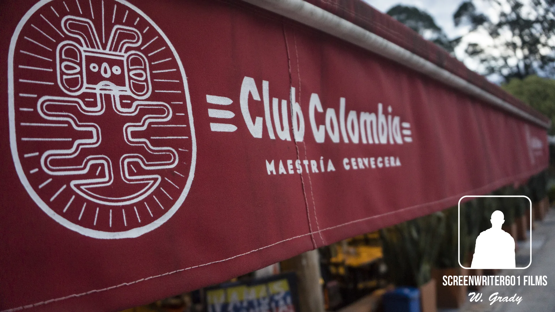 Close view along a dark red awning printed with a circular line-drawn emblem and the words “Club Colombia — Maestría Cervecera”, market stalls blurred beyond it.