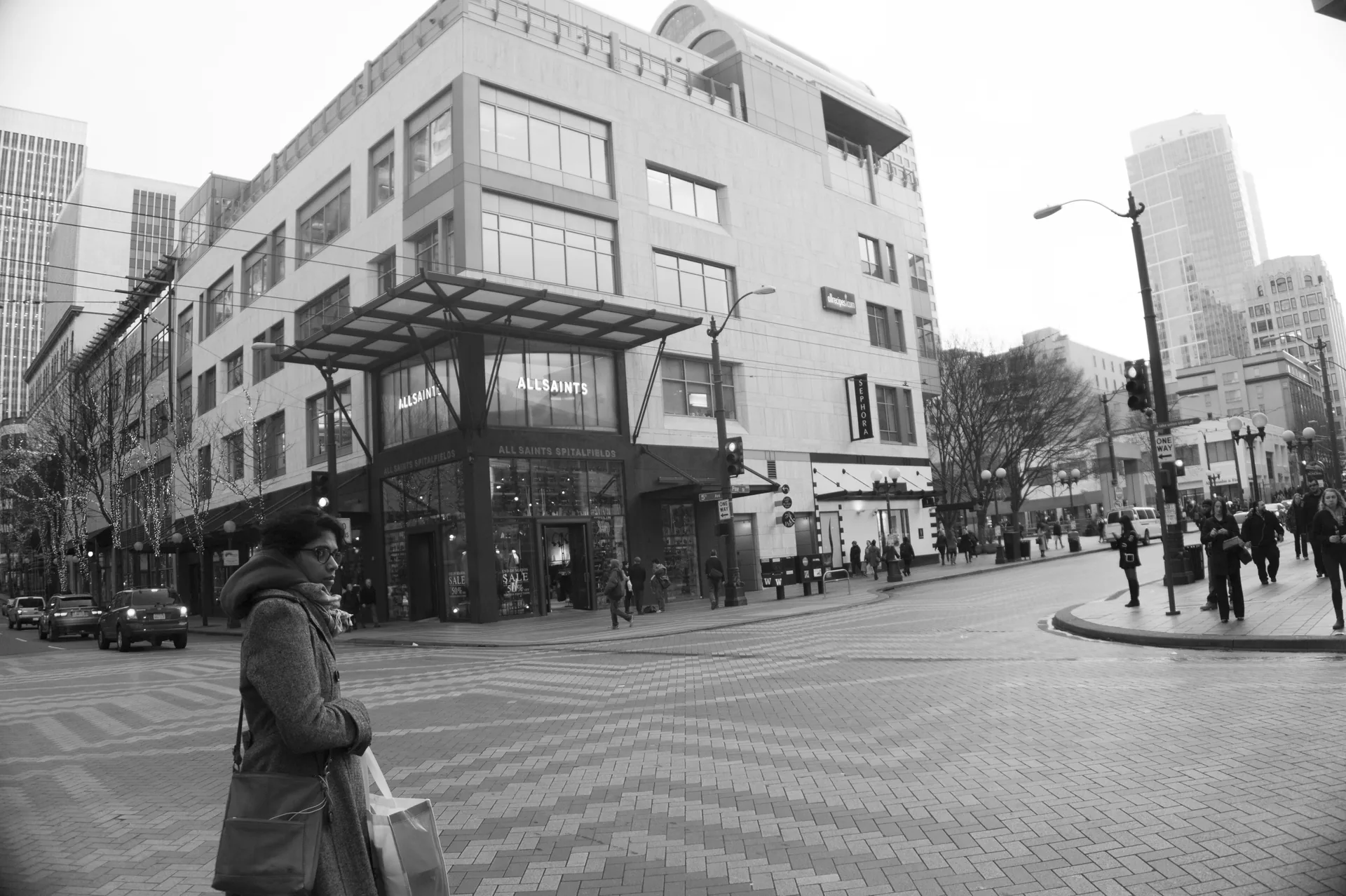 Black-and-white city corner: a woman carrying shopping bags stands in the foreground of a brick plaza before a modern building with an “AllSaints” storefront.