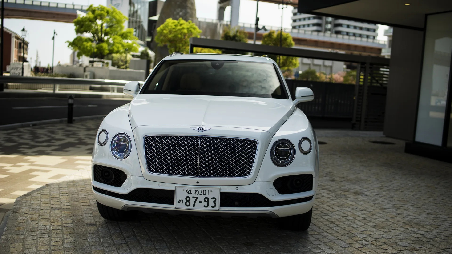 Front view of a white luxury SUV parked on a paved forecourt with a Japanese licence plate, low trees and modern buildings behind it.