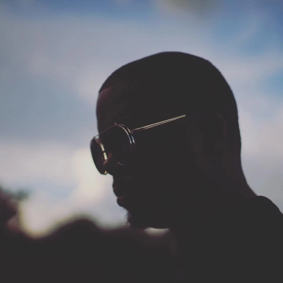 Square front cover of The Most Interesting Nobody: a close, heavily shadowed profile of a person wearing sunglasses, silhouetted against a soft blue and grey sky.