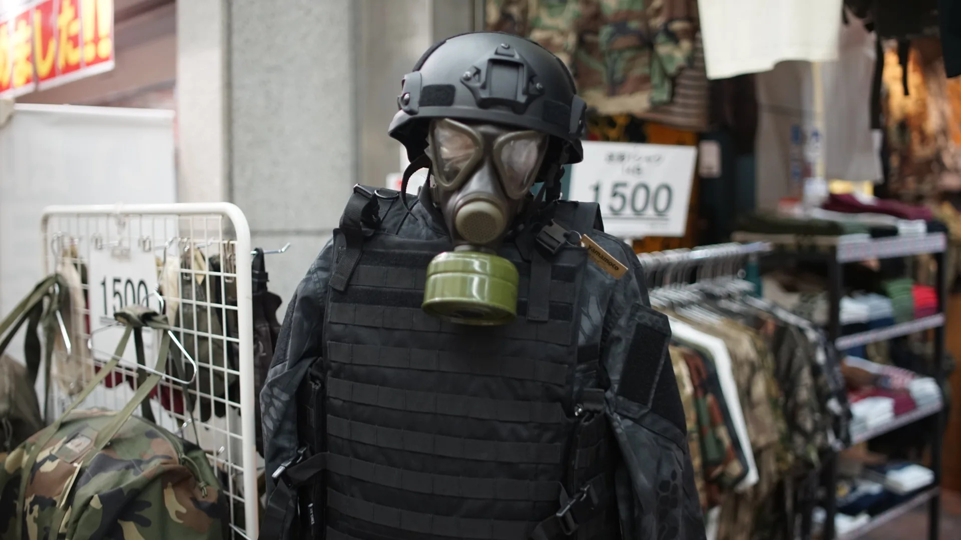 A mannequin displayed outside a shop wearing a black helmet, gas mask and tactical vest, surrounded by camouflage clothing and handwritten price tags.