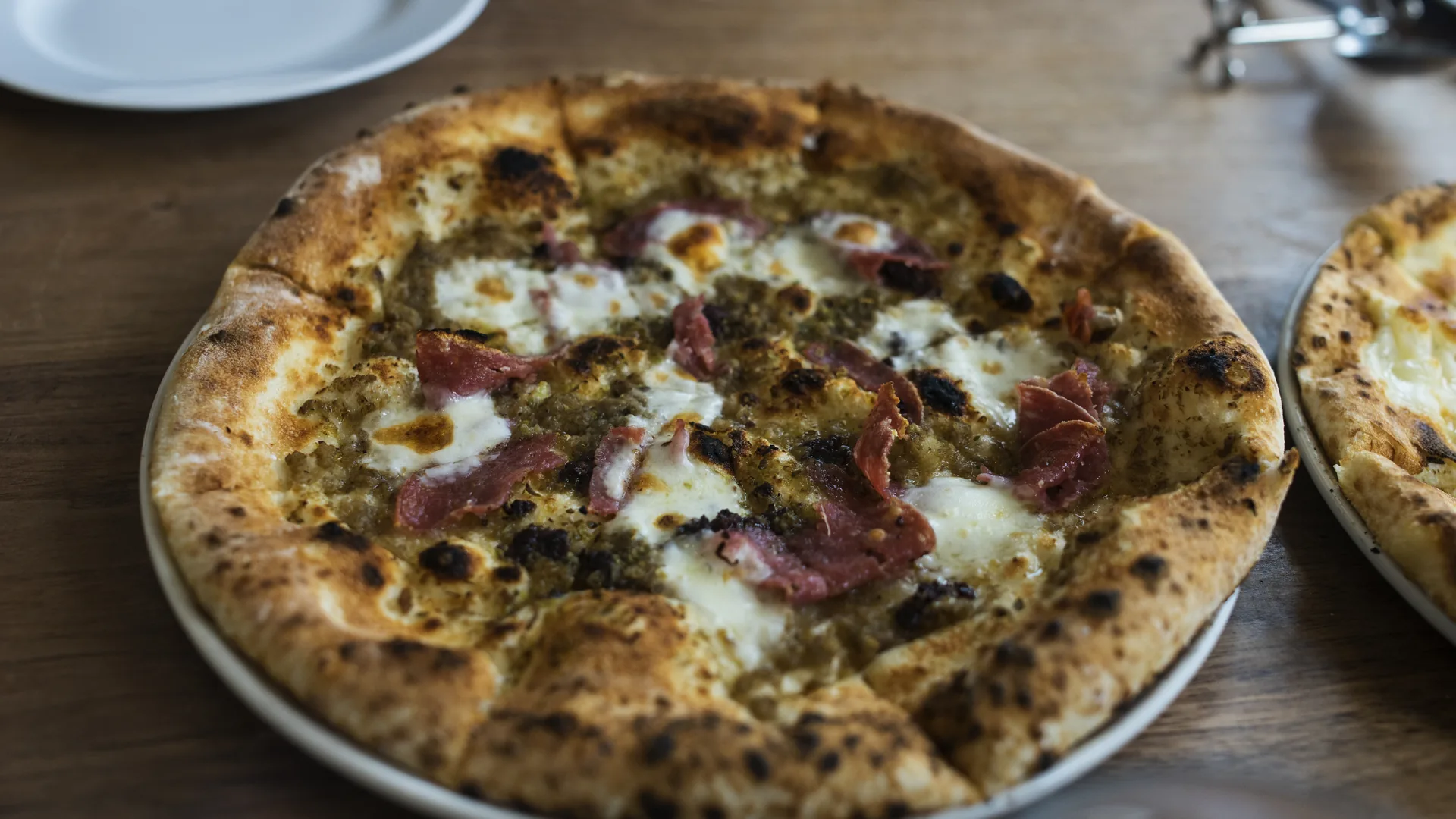 A wood-fired pizza topped with melted mozzarella, green herb sauce and slices of cured meat, resting on a plate on a wooden table with a second pizza at the edge of frame.