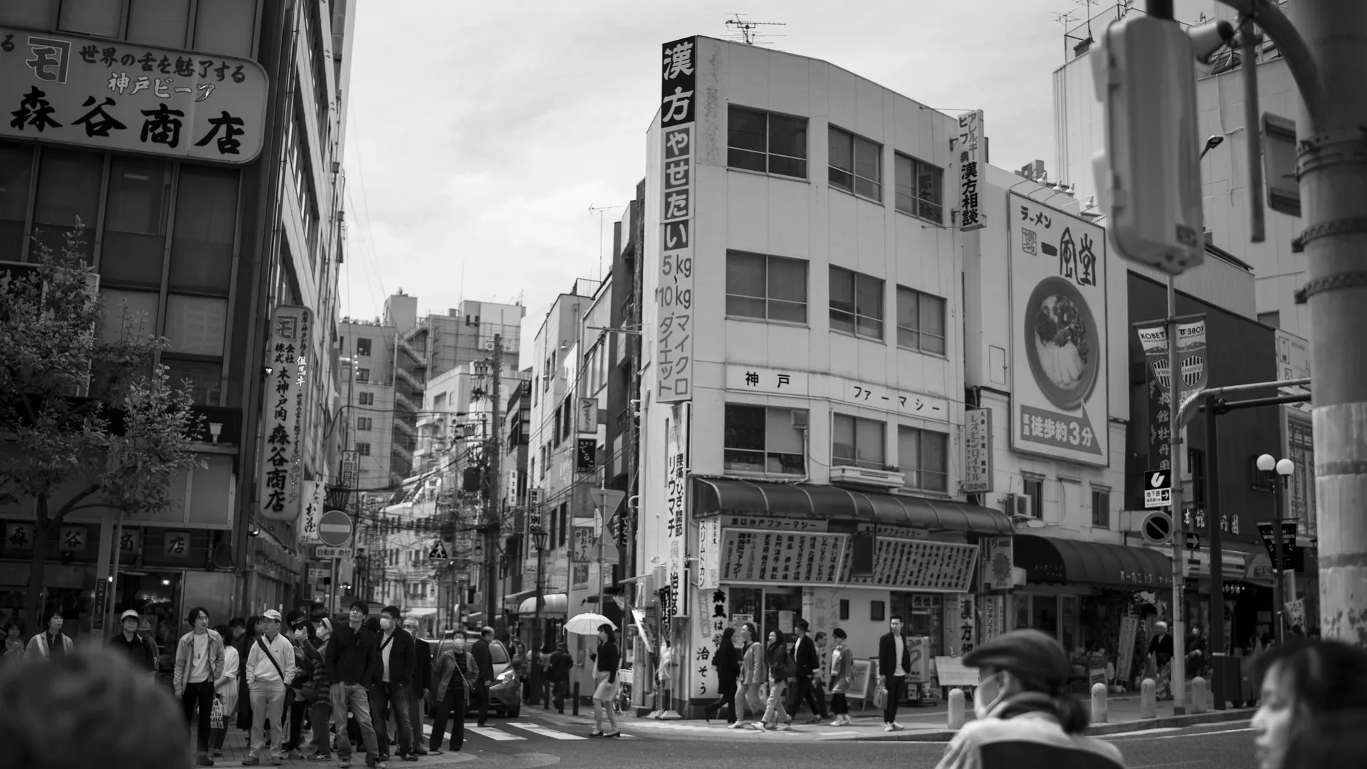 Black-and-white street corner where pedestrians wait to cross, multi-storey buildings behind them covered in vertical Japanese shop signage.