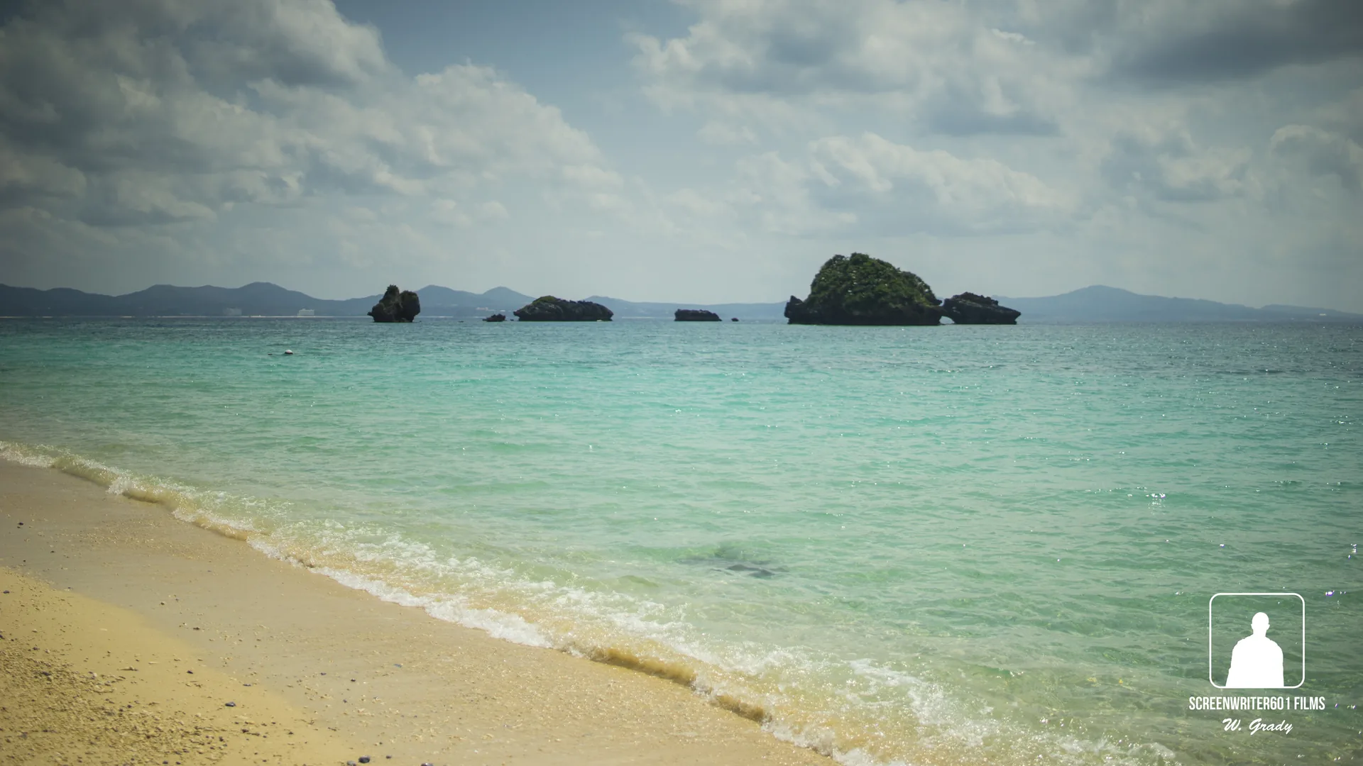 Turquoise shallow sea meeting a pale sand beach, with small rocky islets topped by trees offshore and low hills on the horizon beneath a cloud-filled sky.
