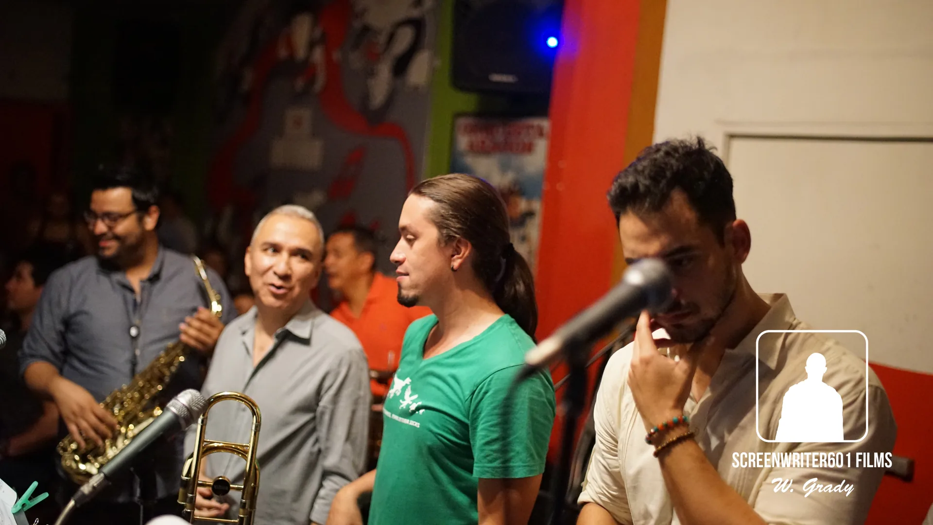 Musicians and onlookers packed together inside a brightly painted bar, a saxophone and a trombone held at the left of frame and a microphone stand in the foreground.