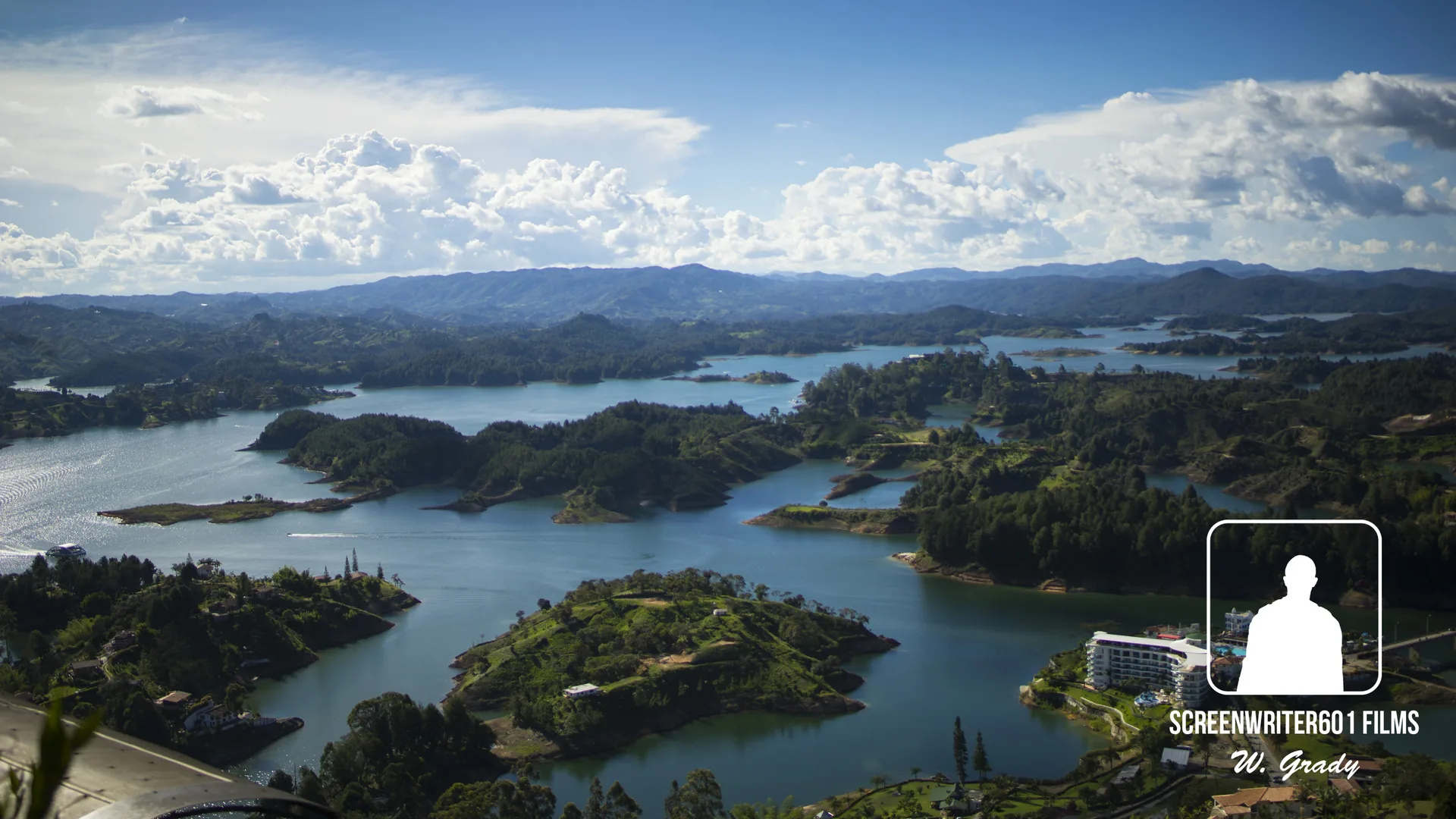 Wide view from above a reservoir dotted with green islands and peninsulas, a large terraced building at the right and mountains along the horizon.