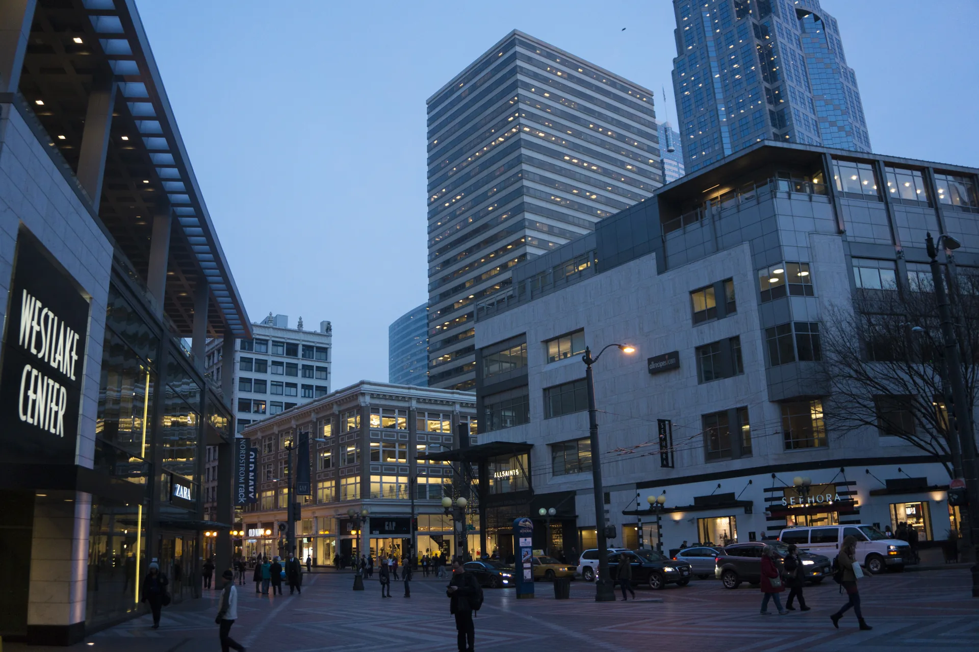 A city square at blue-hour dusk with an illuminated “Westlake Center” sign at the left, lit shopfronts, an office tower behind and people crossing the paved plaza.