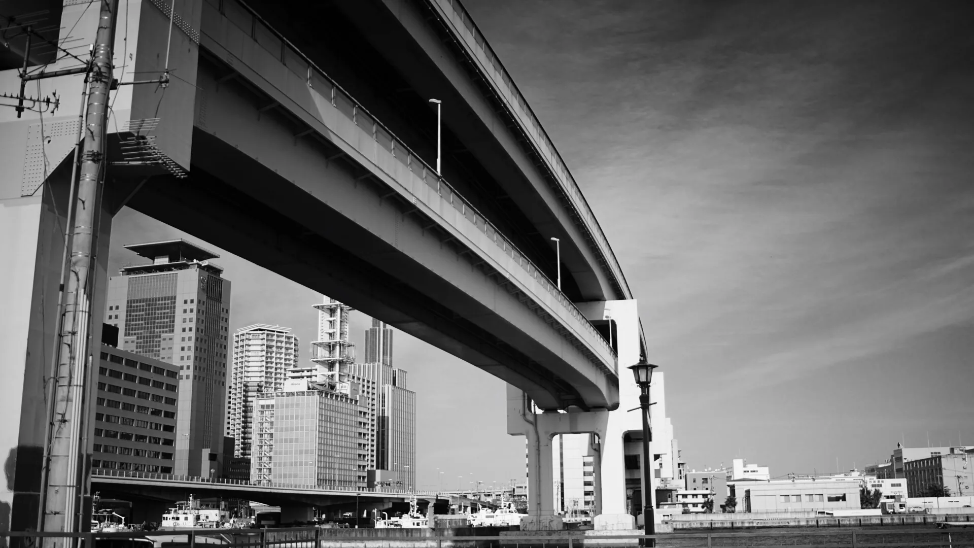 Black-and-white view from below of a curving elevated highway sweeping past mid-rise apartment towers, with boats moored along the waterway underneath.
