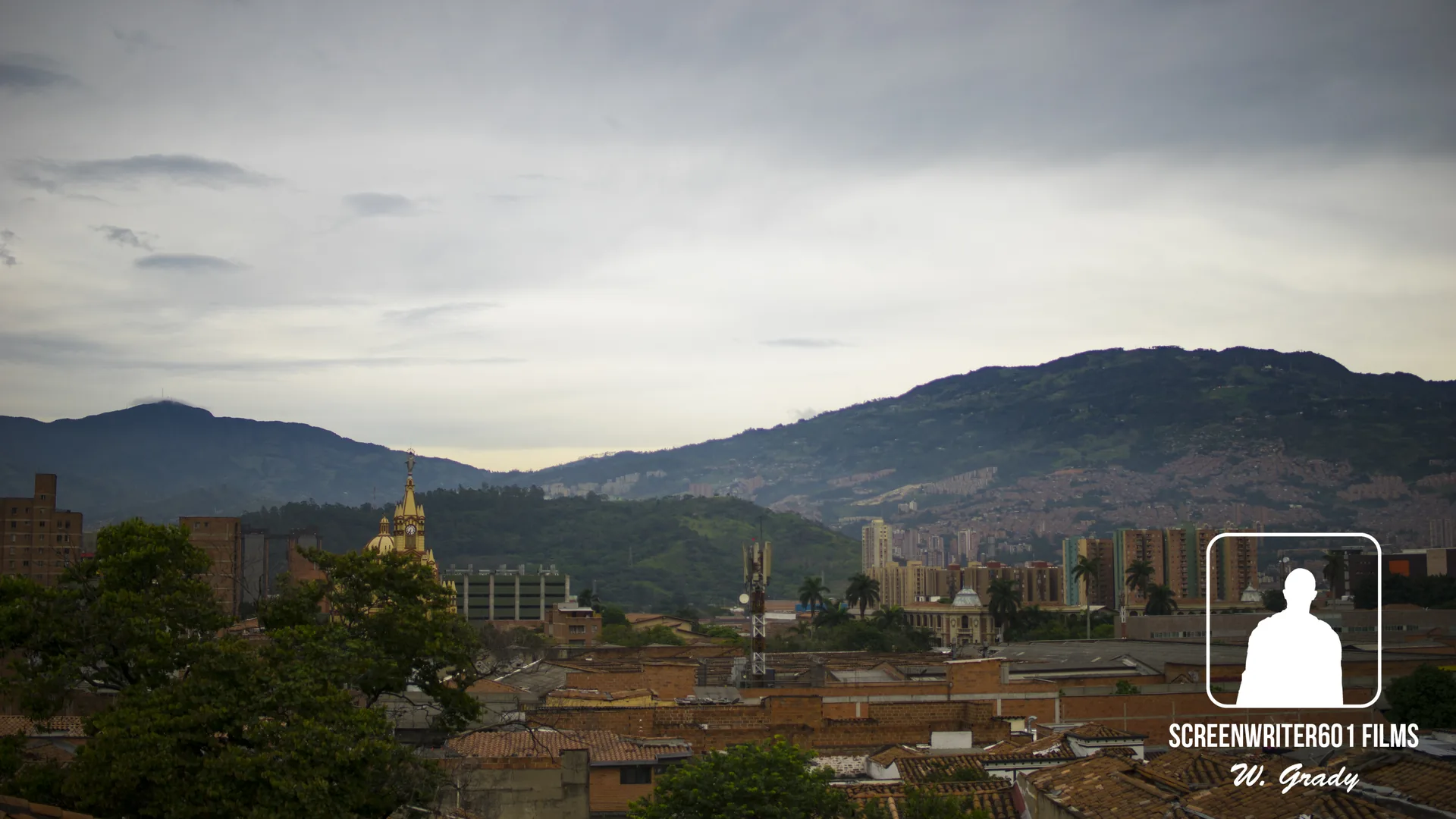 Overcast dusk above a hillside city: tiled rooftops in the foreground, a gilded church spire, apartment towers and communication masts, with mountain ridges behind.