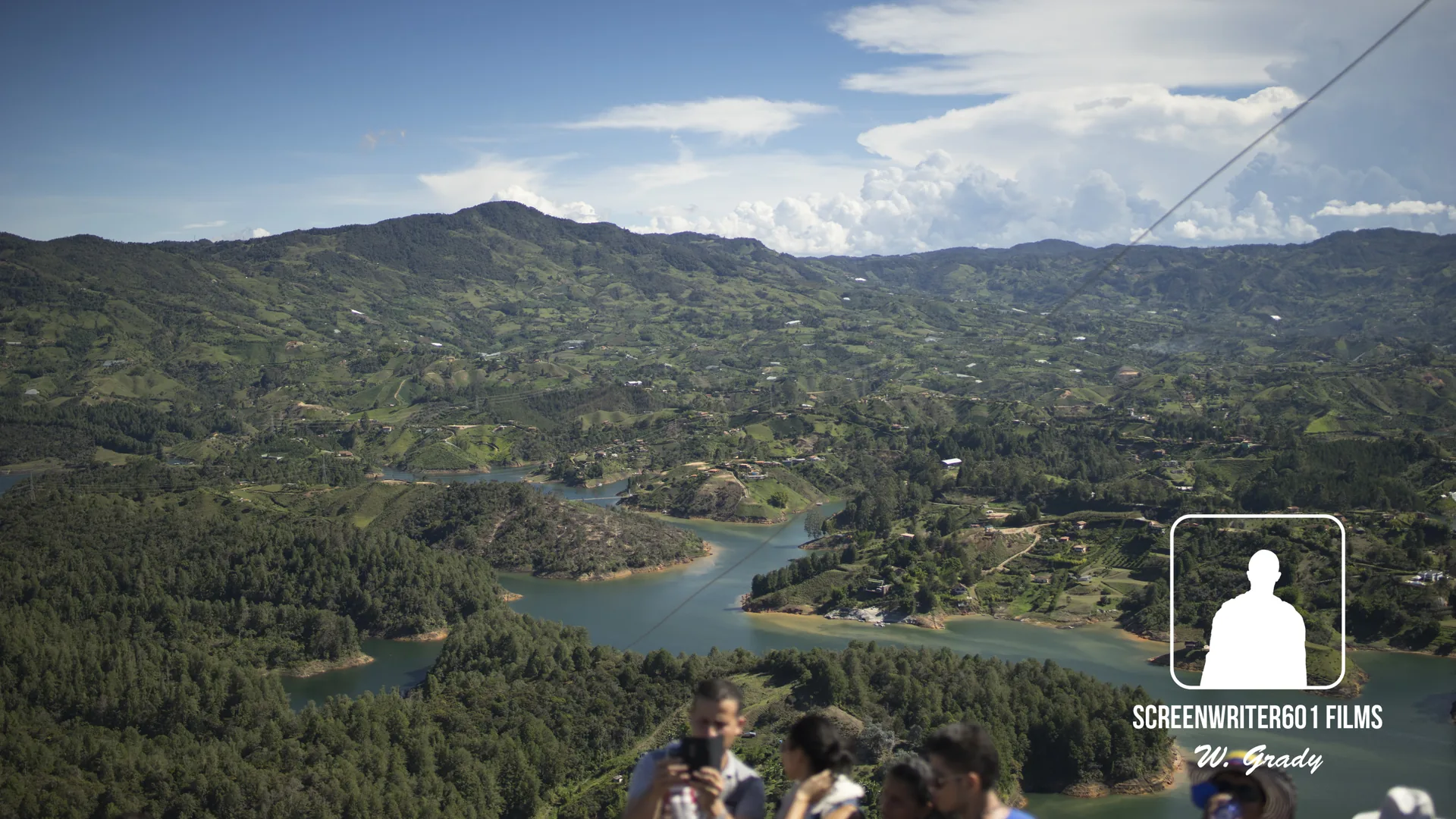 High vantage across forested hills and a winding stretch of water, with visitors at a railing in the foreground raising phones toward the view.