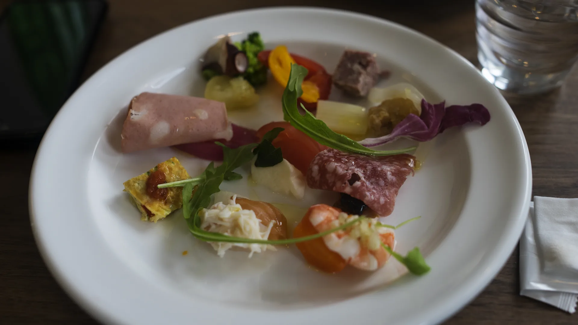 A white plate of small tasting portions — sliced terrine, cured meats, shrimp, shredded crab, pickled vegetables and rocket leaves — on a dark wooden table beside a glass of water.