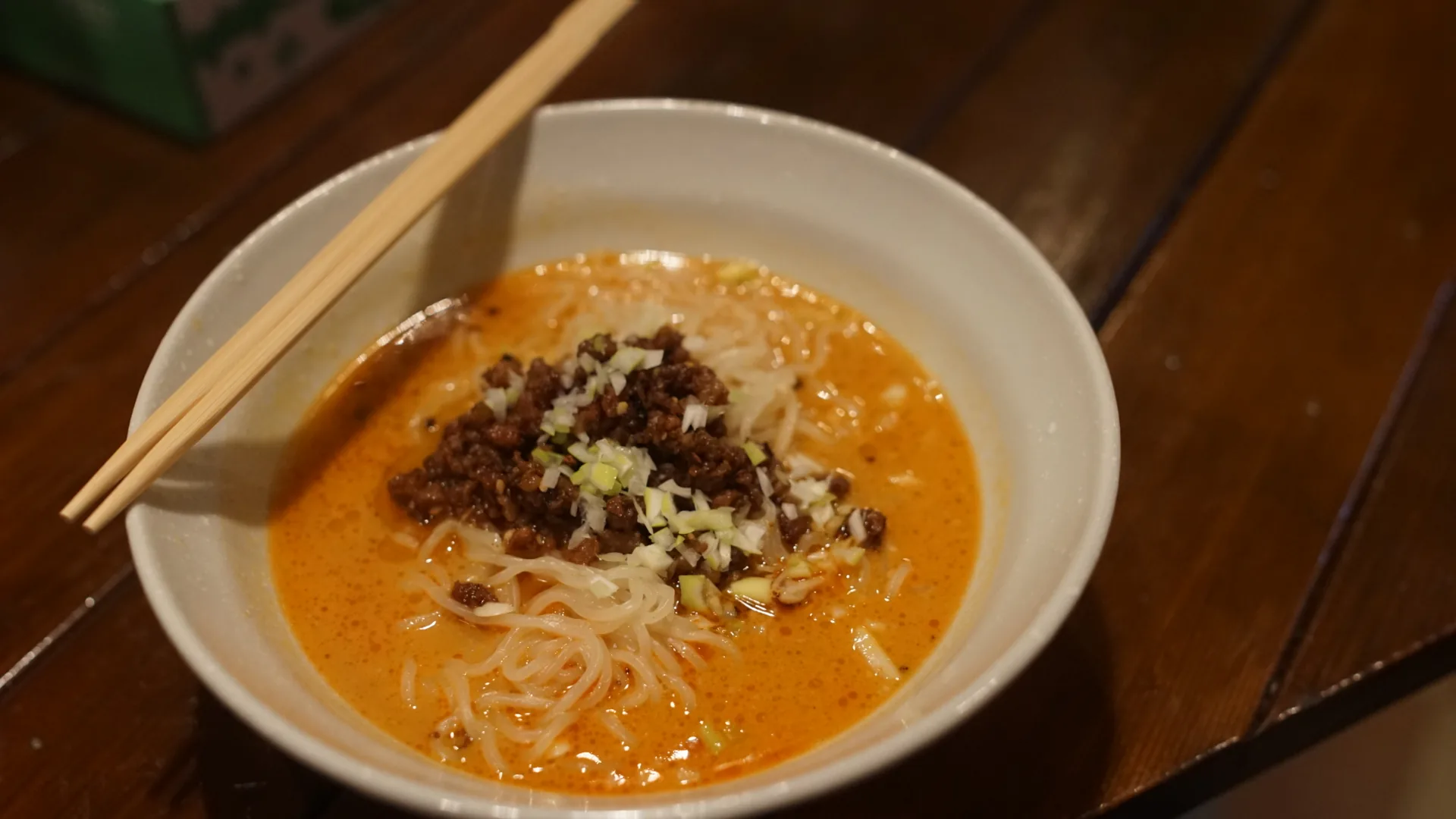 A white bowl of thin noodles in an orange-red sesame broth topped with minced meat and chopped spring onion, wooden chopsticks resting across the rim.