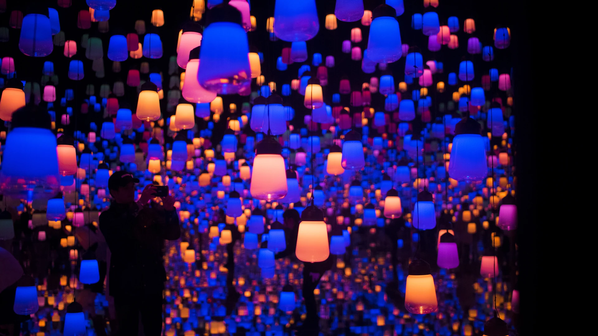 Silhouetted visitors beneath a dense ceiling of glowing lantern-like lights in blue, purple, orange and pink.