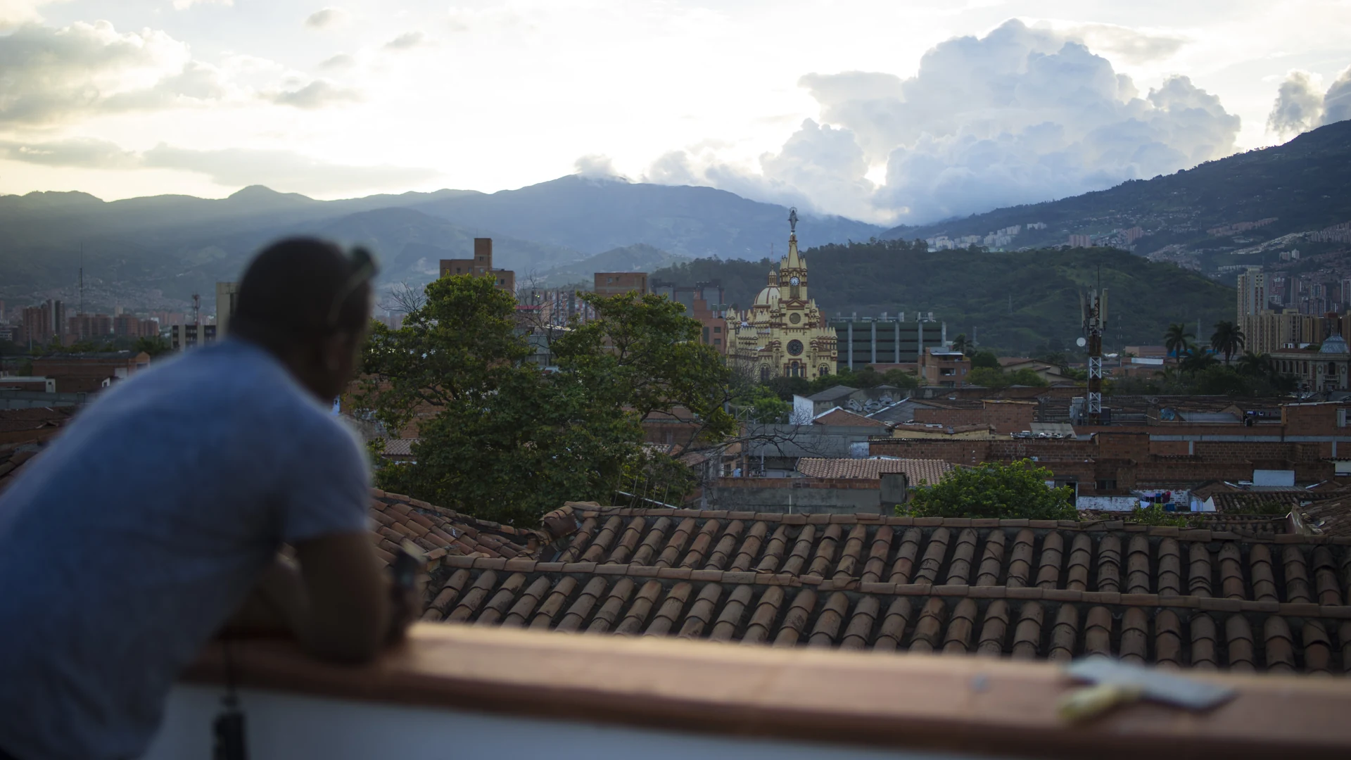 A person seen from behind leans on a terrace railing, looking across a dense city toward cloud-covered mountains.